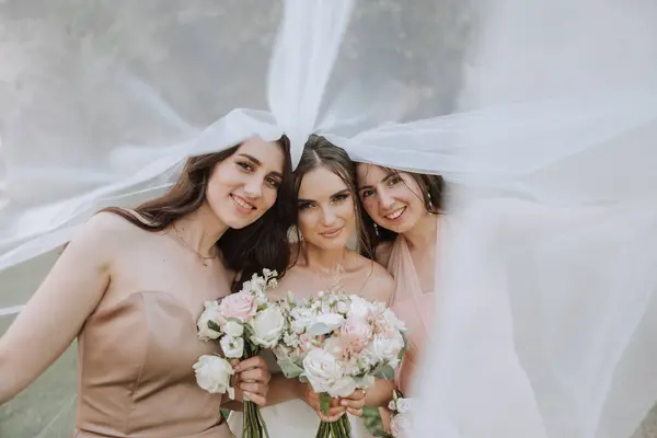 Bridesmaids are wrapped in a veil. The bride is holding a bouquet. Long veil. Wedding in nature. Smiling girls in identical dresses.
