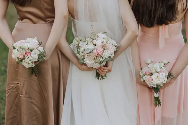The bride and her two friends turned their backs to the camera, holding wedding bouquets of flowers behind their backs.