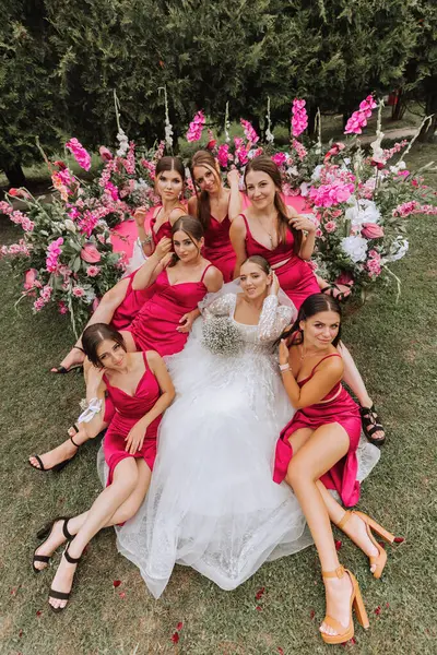 A brunette bride and her bridesmaids in dresses of the same color sit with the bride and rejoice with flowers in their hands near the solemn arch. Wedding in nature.