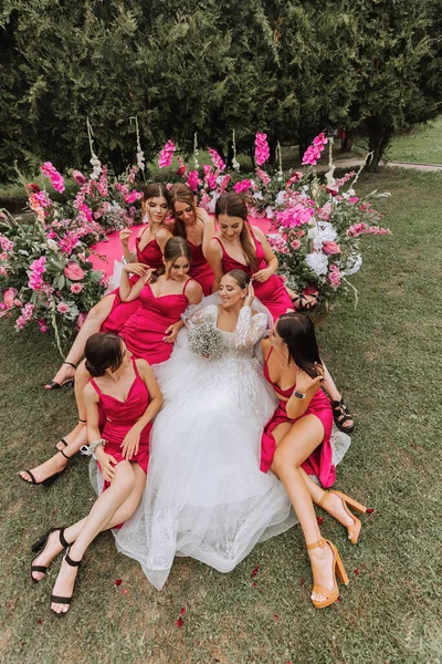 A brunette bride and her bridesmaids in dresses of the same color sit with the bride and rejoice with flowers in their hands near the solemn arch. Wedding in nature.