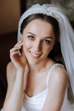 Otel odasında gelinin portresi. Güzel genç bir kız beyaz bir gelinlik giyiyor. Modern düğün saç stili. Doğal makyaj..