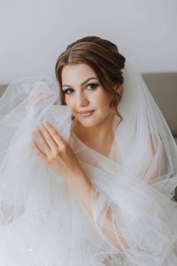 Otel odasında gelinin portresi. Güzel genç bir kız beyaz bir gelinlik giyiyor. Modern düğün saç stili. Doğal makyaj..
