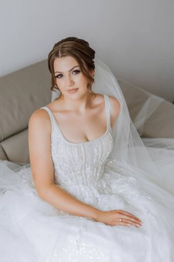 Otel odasında gelinin portresi. Güzel genç bir kız beyaz bir gelinlik giyiyor. Modern düğün saç stili. Doğal makyaj..