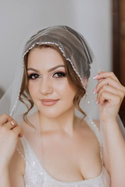 Otel odasında gelinin portresi. Güzel genç bir kız beyaz bir gelinlik giyiyor. Modern düğün saç stili. Doğal makyaj..