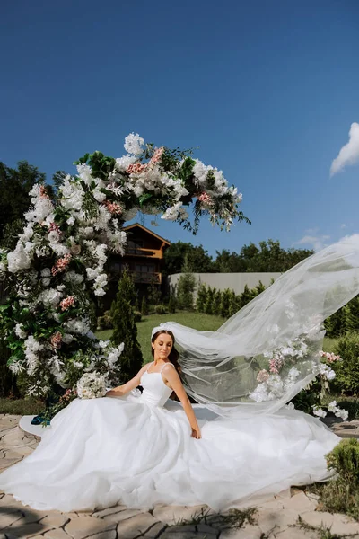A beautiful bride in a white dress with a long train sits near an arch decorated with flowers. The bride in a white dress with a long veil in nature on a summer day. Summer wedding.