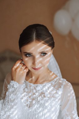 Otel odasında gelinin portresi. Güzel genç bir kız beyaz bir gelinlik giyiyor. Modern düğün saç stili. Doğal makyaj..