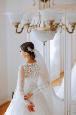 Otel odasında gelinin portresi. Güzel genç bir kız beyaz bir gelinlik giyiyor. Modern düğün saç stili. Doğal makyaj..