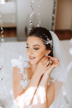 Beyaz dantelli gelinlik içinde güzel, genç, mükemmel bir gelin sabah yatak odasında oturuyor ve kulağına küpe takıyor. Düğün fotoğrafı, portre, konsept.
