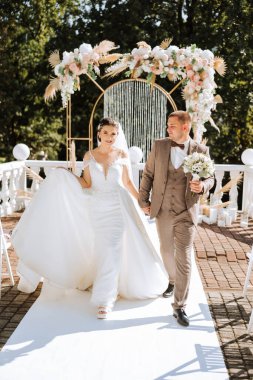 Gelin ve damat düğün kemerinin arka planında yürüyorlar. Açık havada düğün töreni. Damat ve gelin düğün kemerinin arkasında..