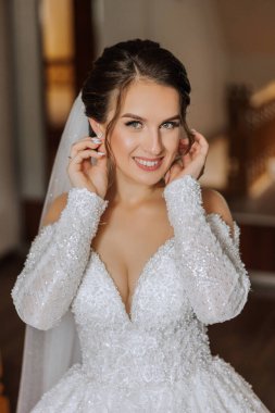 Otel odasında gelinin portresi. Güzel genç bir kız beyaz bir gelinlik giyiyor. Modern düğün saç stili. Doğal makyaj..