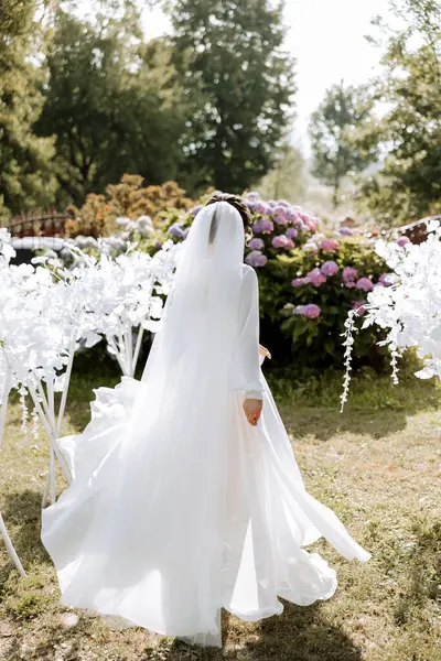 一位穿着雅致长袍的美丽的黑发新娘正在公园里散步 公园里长着绿草和粉红色的水仙花 一个可爱女孩的婚礼肖像 摄影和概念 — 图库照片