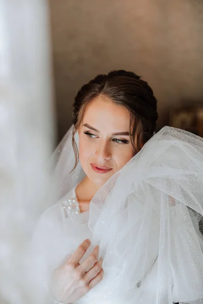 Beautiful bride in a dressing gown in the morning before the wedding ceremony. Incredible hairstyle of the bride. Natural and modern makeup. Portrait of a young bride in a dressing gown.