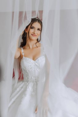 Otel odasında gelinin portresi. Güzel genç bir kız beyaz bir gelinlik giyiyor. Modern düğün saç stili. Doğal makyaj..