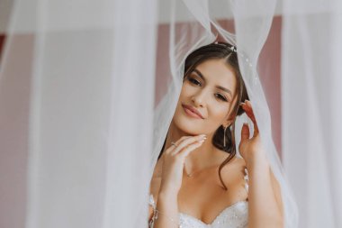 Otel odasında gelinin portresi. Güzel genç bir kız beyaz bir gelinlik giyiyor. Modern düğün saç stili. Doğal makyaj..