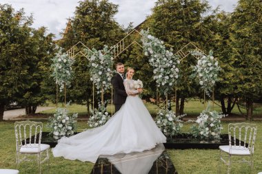 Yakışıklı bir damat, gelinini bembeyaz bir elbiseyle kucaklıyor ve güzel bir açık havada gülümsüyor. Açık gökyüzünün altında. Yüksek kalite fotoğraf. Yeni evli bir çift güneşli bir yaz gününde birlikte poz verir..