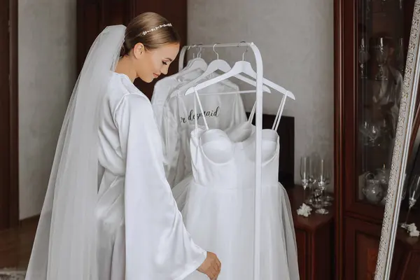 A beautiful bride with a long veil in her room, wearing a robe. Wedding dress on a mannequin. The bride in the morning before the wedding ceremony.