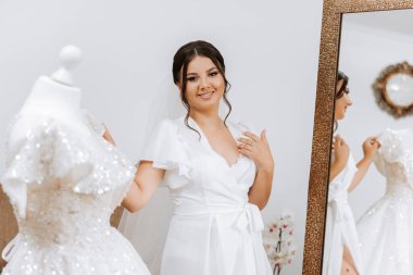 Sabahleyin, inanılmaz güzel ve lüks bir gelin, beyaz bir bornoz giymiş, yatak odasındaki aynanın önünde poz veriyor. Gelinin şaşaalı saç stili ve doğal makyajlı sabahı..