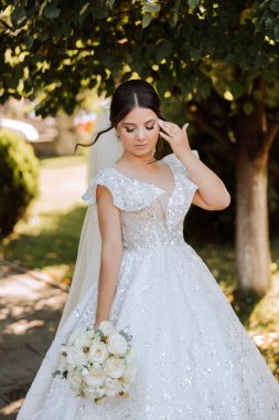 Şehvetli beyaz elbiseli esmer gelin elinde buket, elinde bukleleriyle poz veriyor. Güzel saç ve makyaj. Doğada düğün fotoğrafı çekimi.