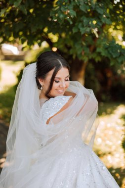 Düğün portresi. Doğada poz veren esmer bir gelin, uzun bir peçeyle örtülü. Güzel makyaj. Dantelli elbise. Düğün töreni.