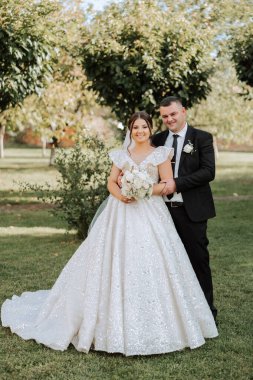 Şık, genç bir damat ve uzun beyaz bir elbise içinde güzel bir gelin ve ellerinde bir buket ile uzun bir duvak, sonbahar doğasında parkta sarılıyorlar. Yeni evlilerin düğün portresi..