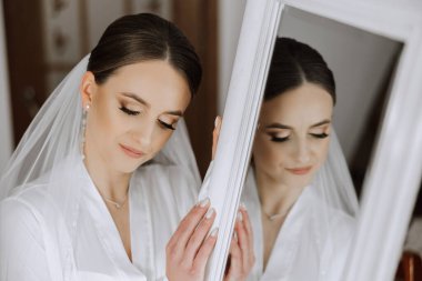 Sabahleyin, inanılmaz güzel ve lüks bir gelin, beyaz bir bornoz giymiş, yatak odasındaki aynanın önünde poz veriyor. Gelinin şaşaalı saç stili ve doğal makyajlı sabahı..