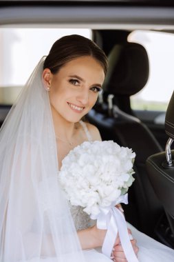 Gelin elinde bir buket çiçekle siyah bir arabada oturuyor ve arabadan dışarı bakıyor. Arabada utangaç bir gelinin portresi. Gelin, gülümse, duygular.