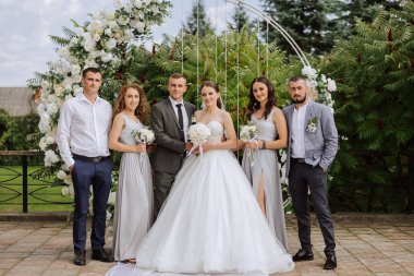 Doğada düğün fotoğrafı çekimi. Gelin ve damat ve arkadaşları çiçeklerle süslenmiş yuvarlak bir kemerin yanında poz veriyorlar. Mutluluk. Bir grup genç insan. Kutlama. Sonbahar düğünü.