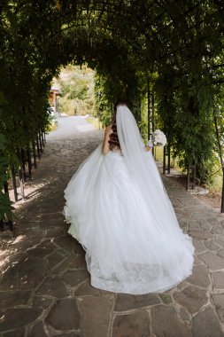 Beyaz elbiseli, uzun trenli esmer gelin elinde buket, bahçede yürüyor. Doğada düğün fotoğrafı çekimi. Güzel saç ve makyaj. Kutlama