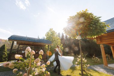 Tarz sahibi damat, kollarında uzun beyaz elbiseler içinde güzel gelini tutar ve sonbahar parkında yeşil çimlerde dans eder, güneş altında orman. Düğün fotoğrafı, gülümseyen yeni evlilerin portresi..