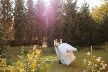 Tarz sahibi damat, kollarında uzun beyaz elbiseler içinde güzel gelini tutar ve sonbahar parkında yeşil çimlerde dans eder, güneş altında orman. Düğün fotoğrafı, gülümseyen yeni evlilerin portresi..