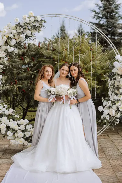 Wedding photography. The bride in a wedding dress and her friends in white dresses pose with bouquets near a white arch. Wedding ceremony. The same clothes. Friendship. Celebration.
