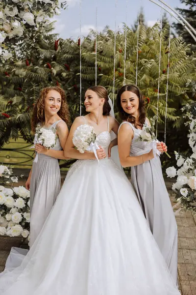 Wedding photography. The bride in a wedding dress and her friends in white dresses pose with bouquets near a white arch. Wedding ceremony. The same clothes. Friendship. Celebration.
