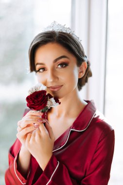 Lüks ve güzel bir gelinin dikey sabah portresi. Kırmızı pijamalarıyla lüks bir odada, elinde kırmızı bir gülle pencerenin önünde duruyor..