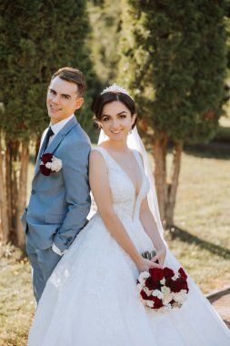 Şık, genç bir damat ve uzun beyaz bir elbise içinde güzel bir gelin ve ellerinde bir buket ile uzun bir duvak, sonbahar doğasında parkta sarılıyorlar. Yeni evlilerin düğün portresi..