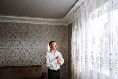 Beyaz gömlekli bir adam odanın penceresinin yanında durur ve yakasındaki ve kollarındaki düğmeleri bağlar. Hazır ol. Bir adamın şık bir iş portresi, yakın çekim fotoğrafı. Damat hazırlanıyor..