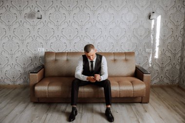 Bir adam deri koltukta oturuyor ve deri kemerli bir saat takıyor şık bir takım elbise, beyaz bir gömlek ve altın bir yüzük takıyor..