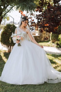 Düğün fotoğrafı. Dantelli, dolgun elbiseli esmer bir gelin elinde bir buket tutarak bahçede yürüyor, içtenlikle gülümsüyor. Harika saç ve makyaj. Moda ve tarz. Kutlama.