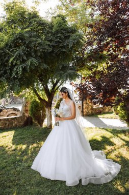 Düğün fotoğrafı. Dantelli, dolgun elbiseli esmer bir gelin elinde bir buket tutarak bahçede yürüyor, içtenlikle gülümsüyor. Harika saç ve makyaj. Moda ve tarz. Kutlama.
