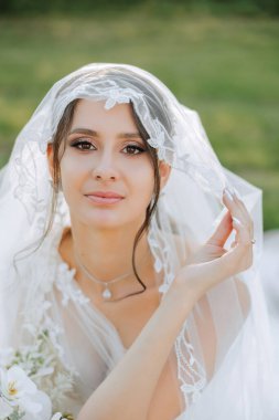 Düğün portresi. Esmer bir gelin elinde bir buket tutuyor ve uzun bir peçeyle doğada poz veriyor. Güzel makyaj. Dantelli elbise.