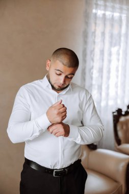 Beyaz gömlekli bir adam odanın penceresinin yanında durur ve yakasındaki ve kollarındaki düğmeleri bağlar. Hazır ol. Bir adamın şık bir iş portresi, yakın çekim fotoğrafı. Damat hazırlanıyor..