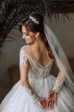 Genç gelin arka tarafta duruyor. Gelinlik giymiş güzel bir kadın, pencerenin yanındaki bir odada. Uzun trenli beyaz gelinlikli çekici kız.