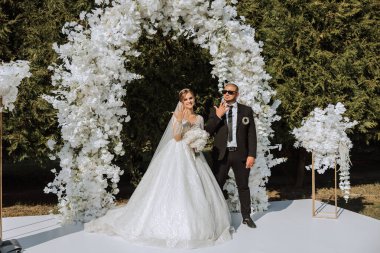 Siyah takım elbiseli şık bir damat ve beyaz dantelli sevimli bir gelin sonbahar doğasının arka planına karşı çiçeklerden yapılmış yuvarlak bir kemer içinde duruyor ve bebek yüzükleriyle gösteriş yapıyorlar..