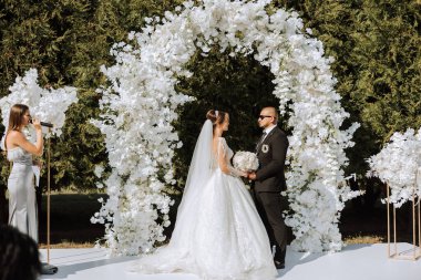 Bir kadın düğün sunucusu konuşma yapıyor ve gelin ve damat düğün töreninde beyaz bir çiçek kemerinin yanında el ele tutuşuyorlar. Gelenekler. Kutlama.
