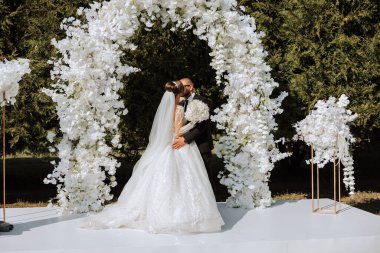 Siyah takım elbiseli şık bir damat ve beyaz dantelli bir gelin sonbahar doğasının arka planında çiçeklerden yapılmış yuvarlak bir kemer içinde durur ve öpüşürler..