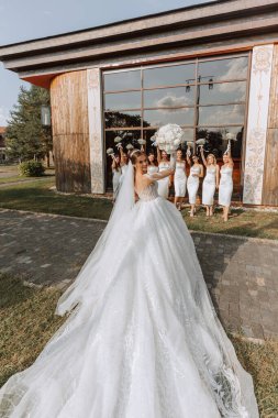 Düğün fotoğrafçılığı. Duvağı olan uzun gelinlikli gelin, beyaz elbiseli arkadaşlarının önünde geniş pencereli bir binanın arka planına karşı poz veriyor. Arkadaşlık. Bir grup genç kız. Kutlama.