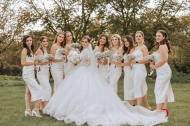 Düğün fotoğrafçılığı. Beyaz bir buketi olan esmer bir gelin ve beyaz elbiseli esmer arkadaşları bahçede bir buket çingene çiçeğiyle poz veriyorlar. Arkadaşlık. Bir grup genç kız. Kutlama.