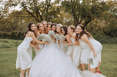 Düğün fotoğrafçılığı. Beyaz bir buketi olan esmer bir gelin ve beyaz elbiseli esmer arkadaşları bahçede bir buket çingene çiçeğiyle poz veriyorlar. Arkadaşlık. Bir grup genç kız. Kutlama.