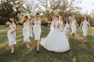 Düğün fotoğrafçılığı. Beyaz elbiseli, buketli esmer bir gelin ve beyaz elbiseli esmer arkadaşları bahçede yürürken poz veriyorlar. Arkadaşlık. Bir grup genç kız. kutlama.