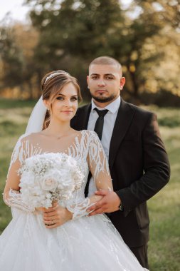 Şık, genç bir damat ve uzun beyaz bir elbise içinde güzel bir gelin ve ellerinde bir buket ile uzun bir duvak, sonbahar doğasında parkta sarılıyorlar. Yeni evlilerin düğün portresi..