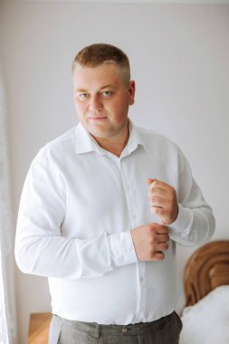 Beyaz gömlekli bir adam odanın penceresinin yanında durur ve yakasındaki ve kollarındaki düğmeleri bağlar. Hazır ol. Bir adamın şık bir iş portresi, yakın çekim fotoğrafı. Damat hazırlanıyor..
